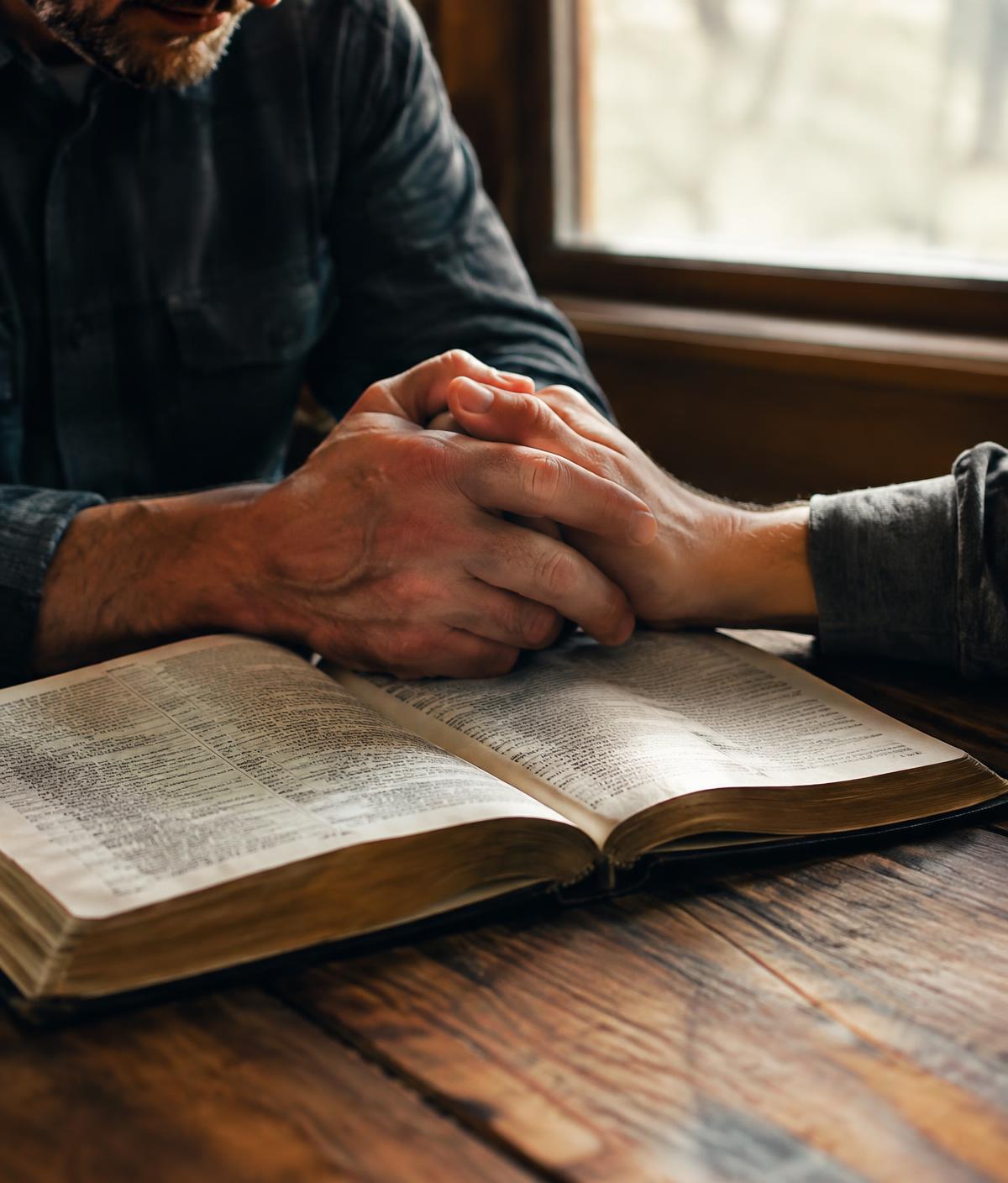 Two men's hands clasped in prayer over an open Bible